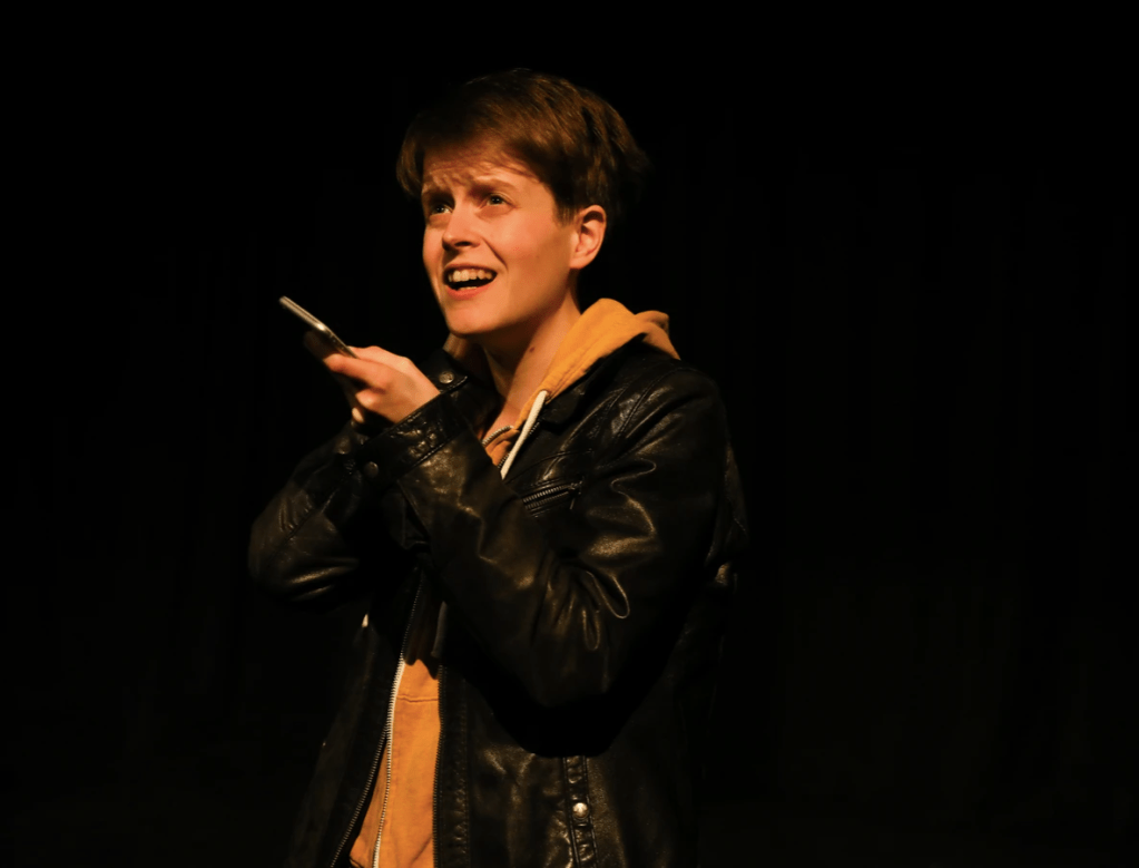 Erin is dressed in a yellow hoody and leather jacket, stood on stage speaking into a phone